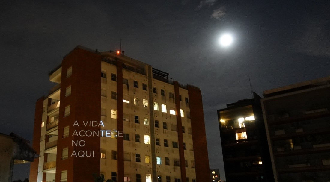 Prédio residencial vermelho e bege, iluminado pela lua, com uma projeção escrito 